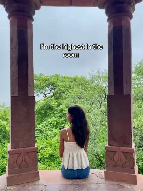 Scenic shot featuring a young woman seated, viewed from behind, within an architectural frame defined by two imposing pillar-like structures. Dense green foliage fills the background under a somewhat overcast sky. Above the woman, overlaid text states, 'I'm the highest in the room'.