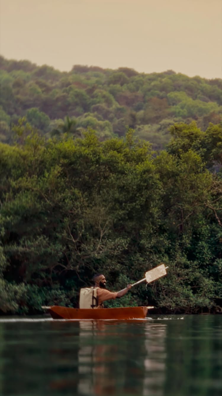 A serene landscape shot of a man in a small boat on calm water. Behind him is a lush green forest and a muted, yellow-toned sky.