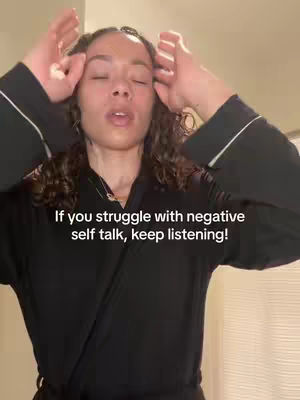 A woman with dark, curly hair stands close to the camera. She is wearing a dark robe with white trim and has her fingers near her forehead. The background is a neutral color.