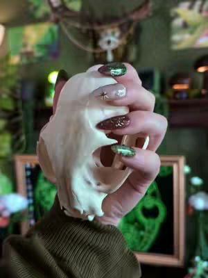 A hand holding a skull and showing off a custom nail set with a thematic design to match the user's gaming setup. The nails feature a variety of colors and details, including green, gold details and woodgrain texture.