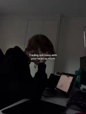 A dimly lit room with a person, obscured by shadow, sitting at a desk with a laptop displaying a trading chart. The person appears to be resting their head on their hand, with overlaid text reading 'Trading will mess with your head so much'.