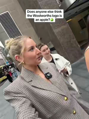 A blonde woman in a plaid blazer is the focus, talking and looking slightly off-camera. A younger woman is visible in the background. The setting appears to be outdoors in an urban environment.