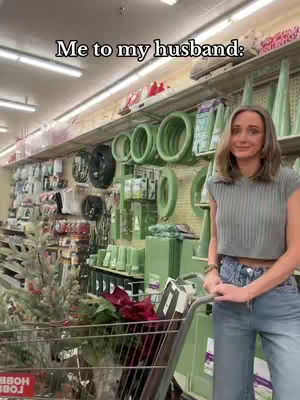 A woman in a green top and jeans stands in a Hobby Lobby store, the background filled with Christmas decorations and shelves of craft supplies. She is standing in front of a shopping cart.