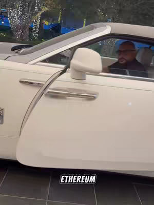 A man in a Rolls-Royce convertible is featured, the car door is open. The image is shot at a low angle, with the car taking up most of the frame. The man is wearing glasses and looking forward, and the lighting suggests a sunny day or bright artificial lights.