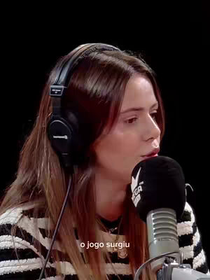 A woman with long brown hair wearing a striped shirt and headphones is speaking into a microphone. The background is black. Text overlayed at the bottom of the image says \"o jogo surgiu\".