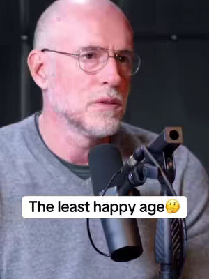 The image is a close-up shot of a middle-aged, bald man wearing glasses and a gray shirt. He is positioned slightly off-center, with a microphone in front of him. The background is dark and out of focus. A text overlay says 'The least happy age' with a thinking face emoji.