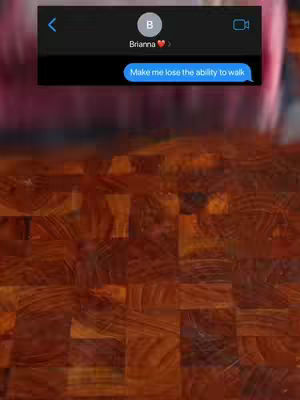 The image shows a screenshot of a text message conversation displayed over a blurred pink and brown background. The text bubble in the message reads "Make me lose the ability to walk". The screen is overlaid onto what appears to be a dark, textured, possibly wooden surface.