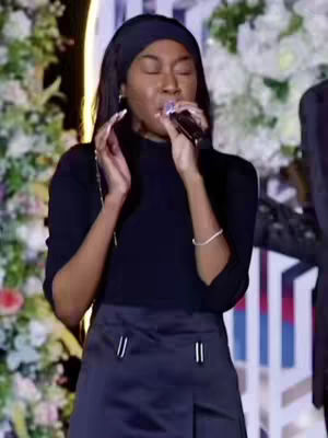 A woman with dark hair and a black headband is singing into a microphone, her eyes closed in what seems to be a worshipful expression. She's wearing a black top and a pleated skirt. The background includes floral arrangements and a pale, vertical striped pattern.