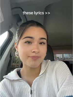 A selfie-style image taken from inside a car. The main subject is a young woman with light skin, dark hair, and a neutral expression. She is wearing a white zip-up sweater and hoop earrings. The text overlay 'these lyrics >>' is near the top of the frame.