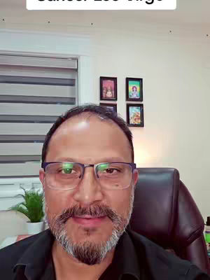 A man with glasses and a beard is centered in a selfie-style video. The background includes a white wall with framed pictures. The man is looking into the camera, with a neutral expression.