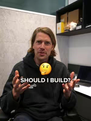 A man with light brown hair is seated, facing the camera. He's wearing a black hoodie, and his hands are up with open palms. An emoji of a thinking face is on his chest overlaid with the text: "SHOULD I BUILD?"