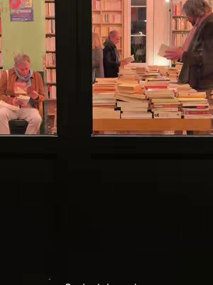 The image captures a bookstore scene through a window. In the left pane, a person sits reading, while in the right pane several individuals browse stacks of books. The scene is warm, with visible rows of books and the interplay of indoor and outdoor light.