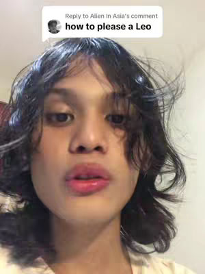 A close-up selfie with a person's face taking up most of the frame. The person has wavy dark hair and is looking towards the camera with a slight pucker of their lips. The background is out of focus and appears to be a neutral color. There is a text box in the upper left that says, 'Reply to Alien In Asia's comment how to please a Leo.'