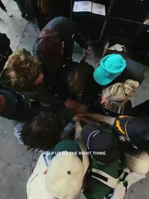 A group of people from a high-angle shot forms a circle, with their hands in the center. Some are wearing casual attire, including caps. The image has a warm, natural lighting.