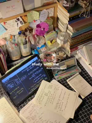 The image shows a workspace filled with study materials, including a tablet displaying handwritten notes, open notebooks, colorful pens, and assorted stationary. Decorative elements such as pink orchids and sticky notes add a visual appeal to the scene, with books stacked in the background.