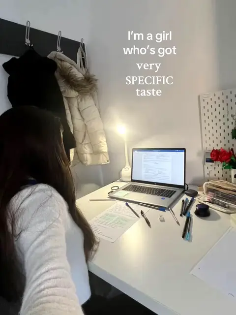 A young woman is seated at a white desk with a laptop open, papers spread out, and various pens and stationary nearby. The desk is against a white wall where coats hang on hooks. A desk lamp is to the right, illuminating the setting. The words "I'm a girl who's got very SPECIFIC taste" are written on the image.