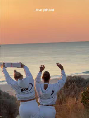 Two young women stand with their backs to the camera, arms raised towards the sky with an ocean and reddish-toned sunset in the background.