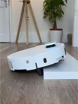 A white robotic vacuum cleaner with a black top is positioned on a wooden floor, seemingly climbing onto a raised platform. A wooden tripod lamp and a potted plant are visible in the background.