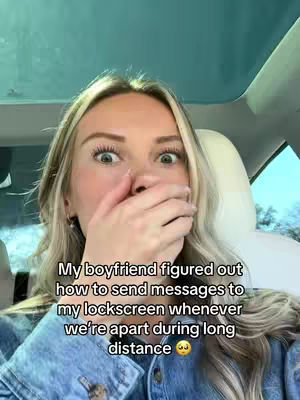 A selfie taken from inside a car features a surprised young woman covering her mouth with her hand. She has blonde hair and wide eyes, with text overlaying the image describing how her boyfriend sends messages to her lockscreen when they are apart.