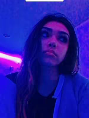 The image features a young woman in a room illuminated with blue and purple lights. She has long dark hair and a serious expression. She is wearing a black shirt and a blue jacket.