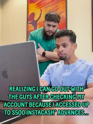 Two men are looking at a laptop screen. One man is sitting in front of the laptop, and the other stands behind him with his arms crossed. The background includes a painting and cream-colored walls.