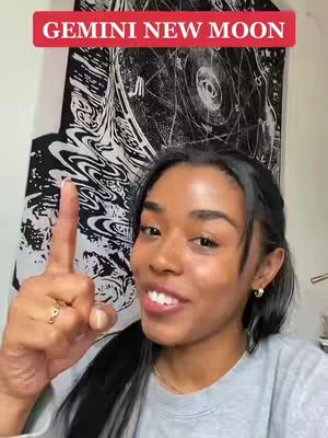 A young Black woman with dark hair looks toward the camera with a smile, holding up her index finger. A red banner at the top of the frame reads, "GEMINI NEW MOON." Behind her is a black and white patterned tapestry.