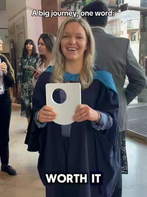 A smiling woman in graduation attire holds a white sign with a circular cutout. Text below the sign reads 'WORTH IT'. Other people are blurred in the background.