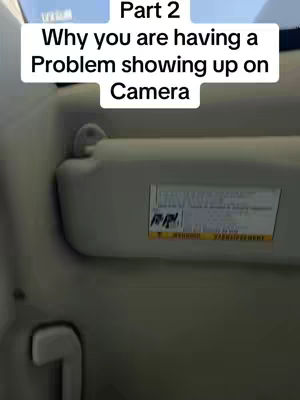 An interior shot of a vehicle’s front seat, with a focus on the upper section. The view is dominated by the interior side of a sun visor, with a label on it. Above the visor, there is a text overlay contained within a white box. The colors are neutral, mainly gray and off-white, with hints of blue through the vehicle's windshield. The composition is straightforward, capturing a moment inside the vehicle.