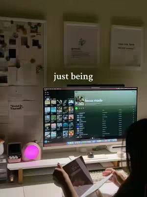 The image showcases a well-lit workspace with a large computer monitor displaying the Spotify interface in focus mode. In the foreground, a person is sitting and reading a book. The desk is organized with a monitor stand, keyboard, and a desk lamp. The background includes notes and pictures on the wall, and two framed pictures. The overall feel is cozy and study-focused.