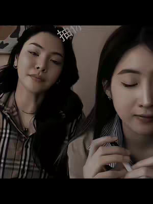 The cover image features two young adult women, captured in a medium close-up. The woman on the left has dark hair styled with a side part, a subtle smile, and is looking towards the camera, wearing a cream-colored blouse and a patterned top, She appears to be in a well-lit indoor setting, with the right half of a second woman visible, with dark hair and is looking down. The color palette leans towards warm tones, with blacks and creams providing balance.