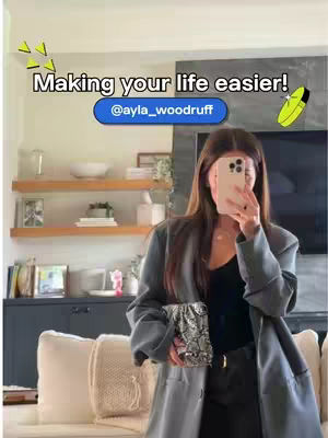 A woman with long brown hair holds a phone up to her face, partially obscuring it. She is wearing a grey blazer over a black top and black pants. She carries a small, patterned clutch. The background includes shelves with decor and a couch.