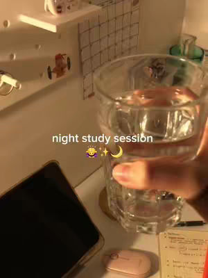 An eye-level shot indoors, featuring a person's hand holding a clear glass mostly filled with water. The background includes a shelf with items, part of a calendar, and a table with a phone, a computer mouse, and some notes. The lighting is warm, creating a cozy ambiance.
