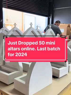 The image shows a workshop setting. In the foreground, several white, altar-like structures with crosses on top are arranged. In the background, a person works near an open doorway, with some cardboard boxes visible.
