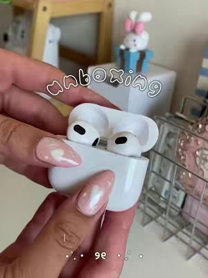 A person's hands holding an open white AirPods case, with the earbuds visible inside. The person's nails are painted with a light pink color, and a heart design can be seen on some nails. In the background, there are other decorative items on a white surface, with the word "unboxing" overlaid on the image in a handwritten font.