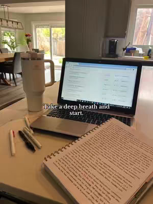 A laptop with text on the screen is open on a desk. A notebook, pens, and a water cup are also visible. The background shows a well decorated kitchen and a door to the outside.
