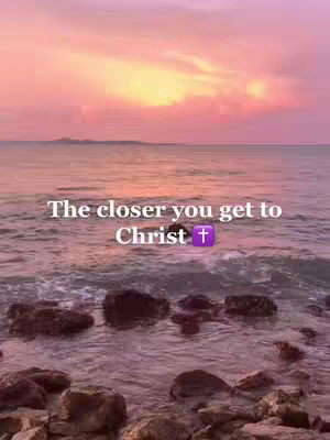 The image captures a scenic view of a beach. The sky is painted with hues of pink and orange during sunset, which creates a vibrant backdrop. The ocean water reflects the sky's colors. In the foreground, dark rocks are partially submerged in the water. Text overlayed reads: 'The closer you get to Christ' with a purple cross emoji.