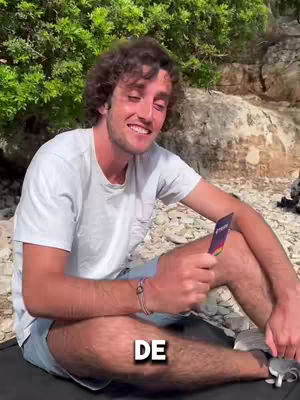 A man is sitting on the ground in front of bushes and rocks, holding a card and smiling. The text "DE" is overlaid on the image.