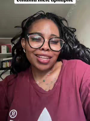 A smiling woman with glasses and long dark hair is centered in the frame, wearing a burgundy t-shirt. Background includes a bookshelf. The composition is a close-up portrait.