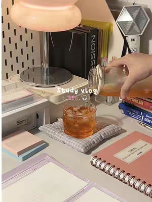 The cover image presents a well-lit, aesthetically pleasing study setup. A clear glass of orange liquid with ice cubes sits on a small, checkered pillow. The scene is captured from a slightly above angle, with soft, warm lighting emanating from a desk lamp and various study materials and decorations. The color palette leans towards soft pastels and neutral tones.