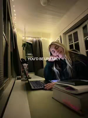 A person, presumably the creator, is shown with long blonde hair, slumped over a laptop on a desk. The scene takes place in a room with warm overhead lighting and fairy lights along the ceiling. A water bottle and books are visible, suggesting a study or workspace.