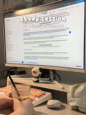 A person's hands are holding a glass of something amber-colored, in the foreground of a desk setup for studying. A computer monitor displays text, including the words ''study session w/ a bunch of research.'' A keyboard and a white mouse are also in the shot, as well as a small panda figurine.