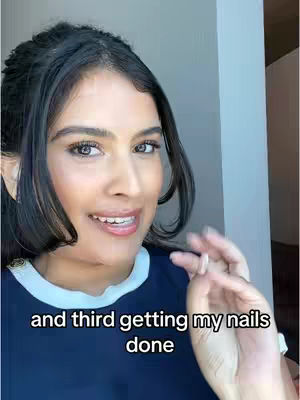 A woman with dark hair, styled away from her face, is centered in the frame. She smiles and looks toward the left. The text overlay reads, 'and third getting my nails done.' She wears a dark blue top with a white collar. The background is a neutral interior.