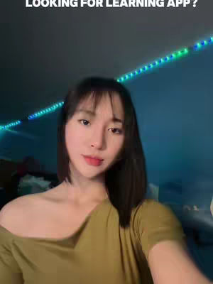 A woman is captured in a selfie from the chest up. She has dark hair with bangs, is wearing a light-colored top, and has red lips. There's a background of blurred lighting, consisting of green and blue tones. The overall effect is casual and inviting.