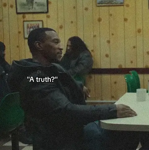 A man in a dark jacket, seated at a table, looks to the left with a thoughtful expression. The background includes a diner-like interior with wood paneling, two additional people are also visible in the background. A text overlay reads "A truth?"