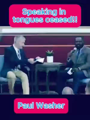 Two men are seated on a stage, one on the left side holding a paper and the other on the right side holding a microphone. There is a small table with a plant between the two men. Text overlays are present at the top and bottom.