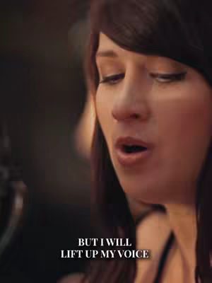 Close-up shot of a woman with dark hair and fair skin, her eyes are closed and her mouth is open as if she is singing or speaking passionately. Below her, overlaid text reads, "BUT I WILL LIFT UP MY VOICE". The background is blurred, suggesting a soft lighting environment that enhances the focus on the woman.