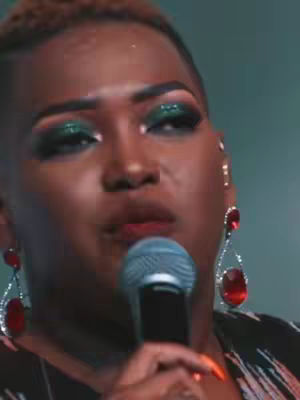 A close-up shot of a person with dark skin wearing heavy makeup, including green eyeshadow and red dangling earrings, holding a microphone. The person appears to be singing or speaking, with a focused expression. The background is blurred, suggesting a performance setting.