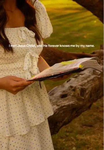 A woman in a white dress holding a book, standing outdoors with a blurred background of greenery and a tree trunk. The lighting is warm and golden.