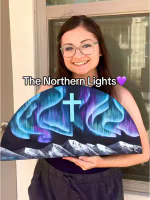 A woman smiles, holding a painting of the Northern Lights with a cross superimposed. The background features a window.