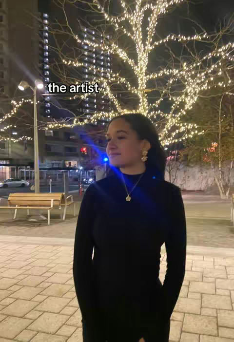 A woman stands on a brick path at night with trees and streetlights in the background. She wears a black top and gold jewelry. The trees are lit with string lights and a sign that says 'the artist'.
